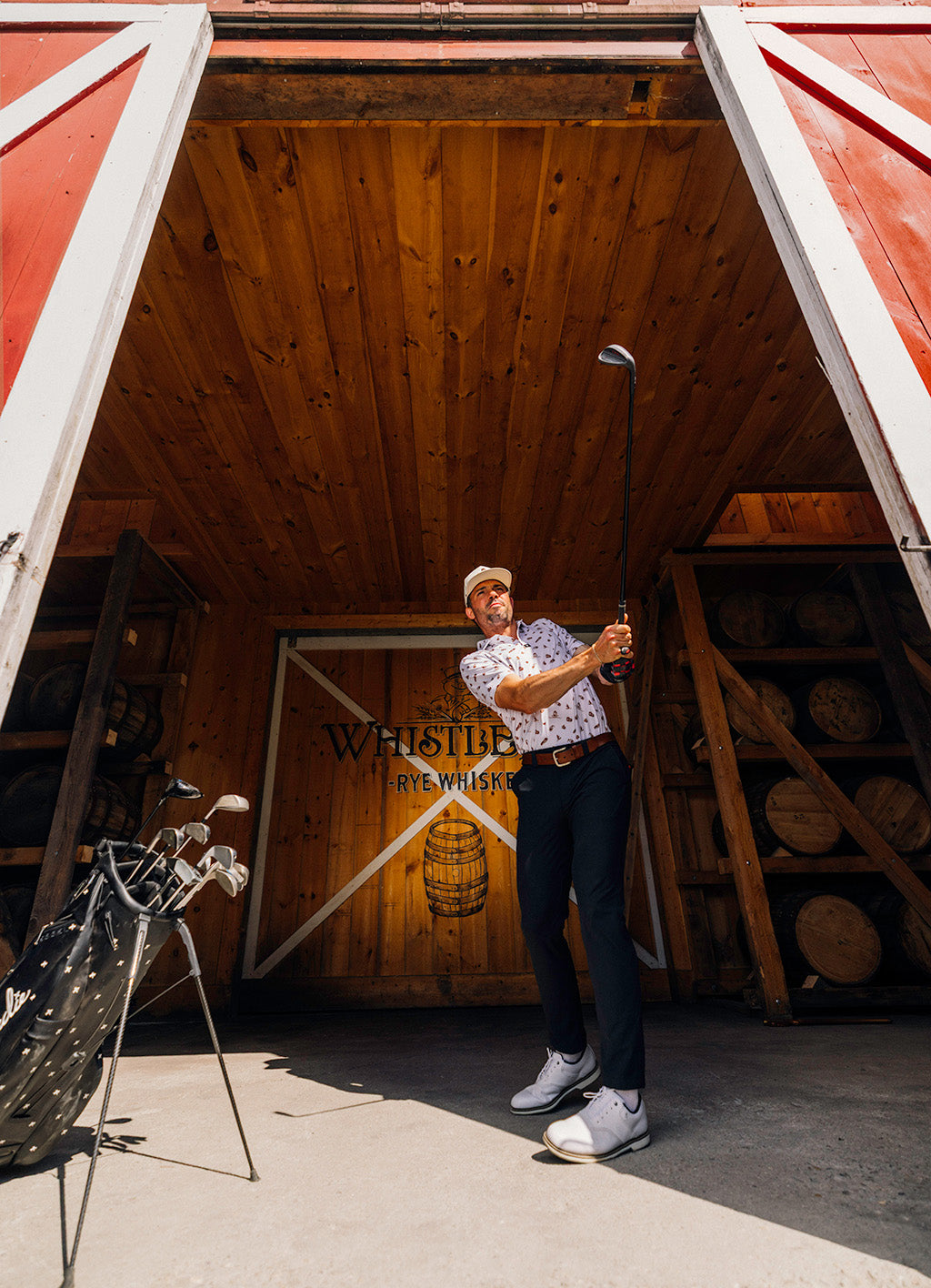 Man holding a golf club in a rustic setting with wooden walls and barrels.